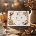A printed invitation with a white background and a border of autumn leaves, pumpkins, and grapes, with text that reads "Save the Date for Friendsgiving Dinner, November 26, 2027, The Smith House," resting on a wooden surface next to a pumpkin and a