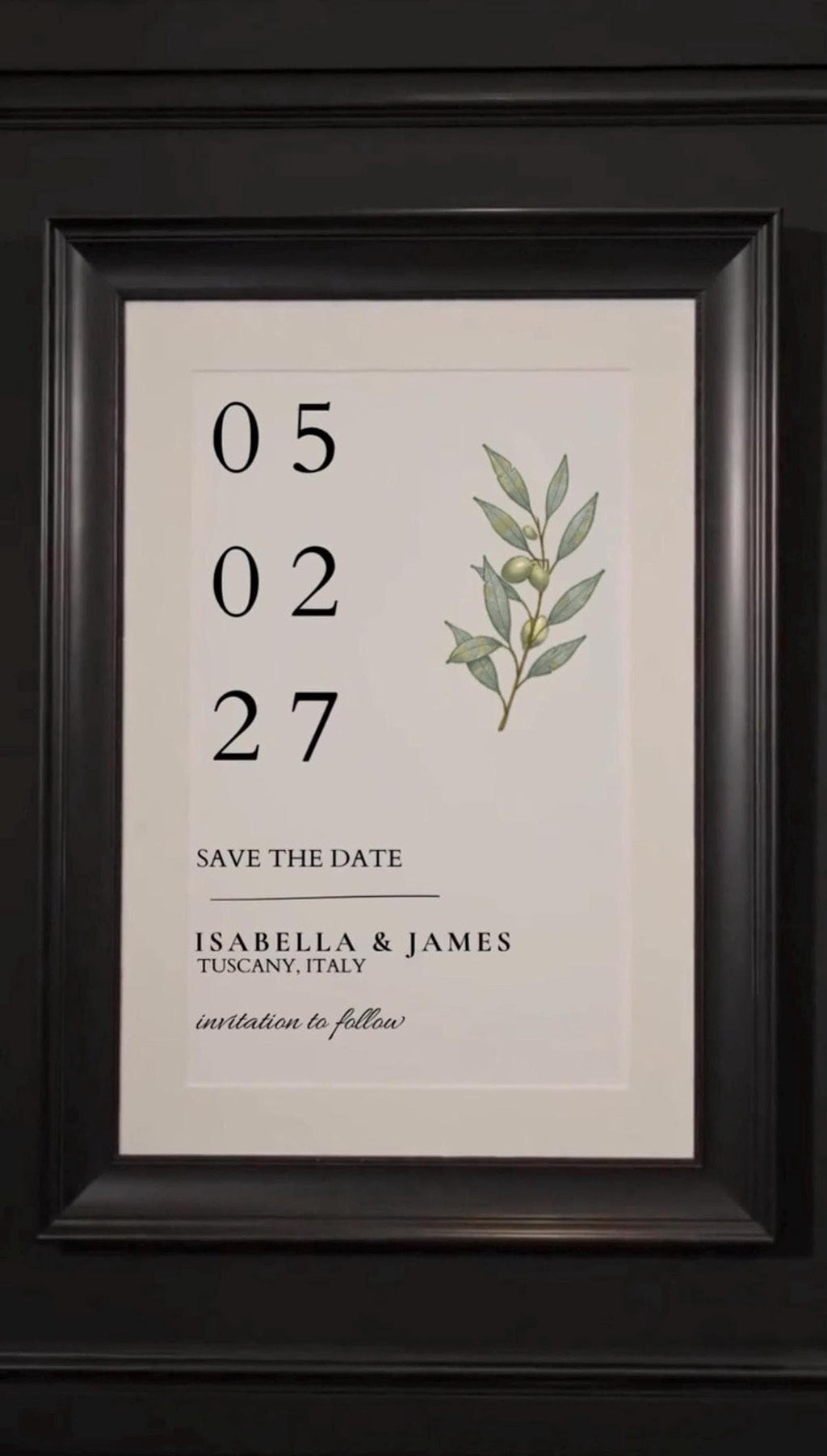 A framed save-the-date card with the date 05/02/27, names Isabella & James, and an olive branch illustration, is displayed on a dark wall.