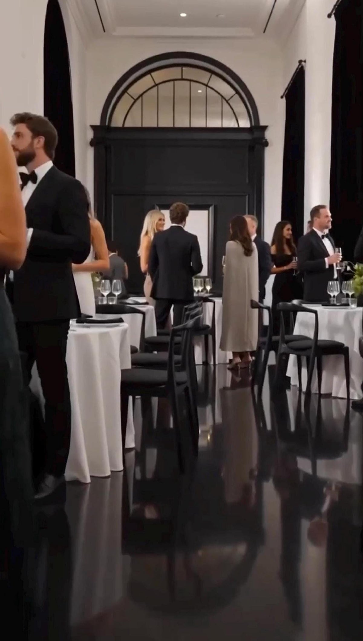 Guests in formal attire mingle in a dimly lit, elegant room with white-clothed tables and dark chairs.
