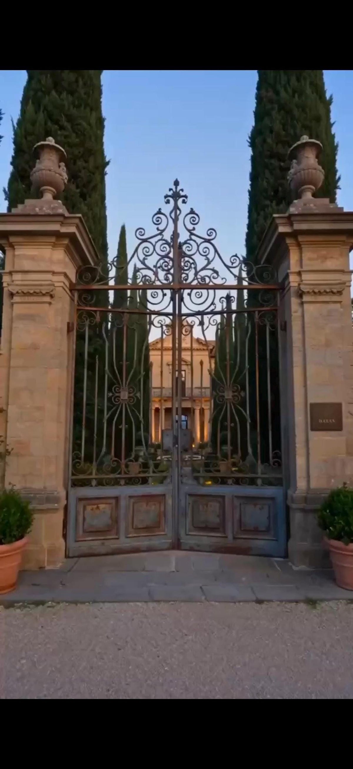 A large, ornate metal gate with stone pillars and cypress trees leads to a distant building.