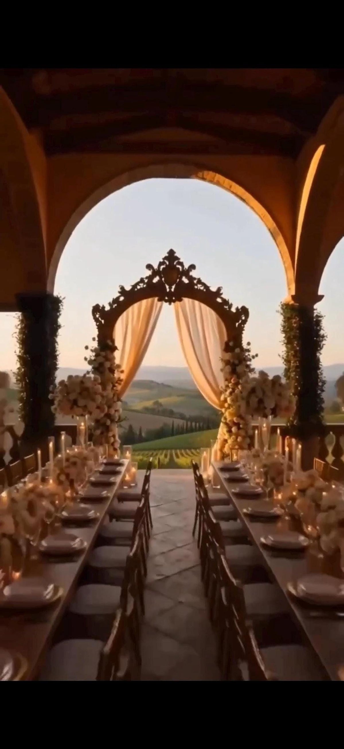 A long dining table set with candles and flowers under an arched canopy, overlooking a vineyard at sunset.