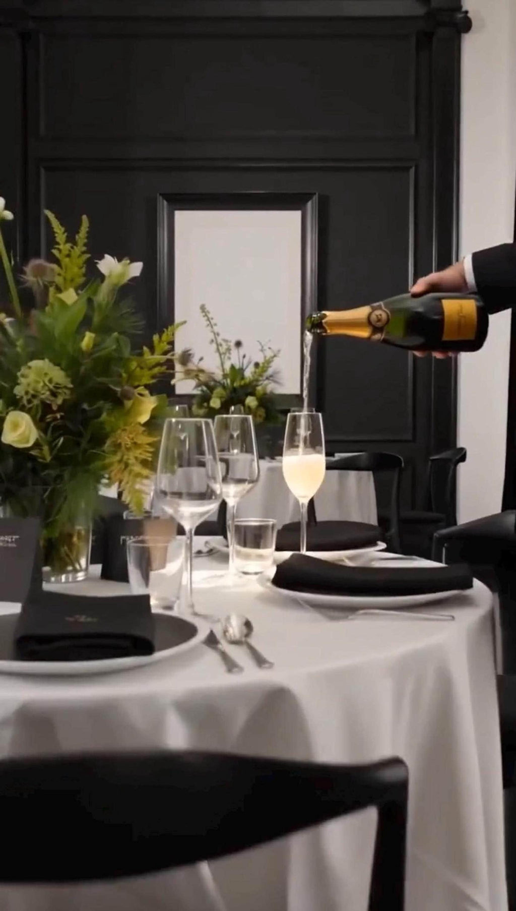 A hand pours champagne into a glass at a formal table setting with a white tablecloth, black napkins, and a floral centerpiece.