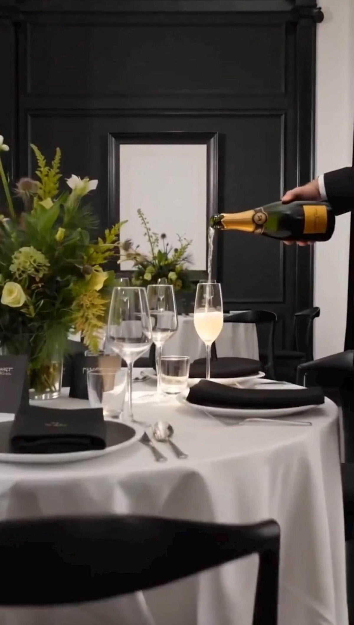 A hand pours champagne into a glass at a formal table setting with a white tablecloth, black napkins, and a floral centerpiece.