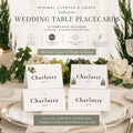 Four white folded place cards are arranged on a white tablecloth, with two featuring a small cluster of grapes and two featuring two tall, slender cypress trees, all reading "Charlotte Smith" in black text.
